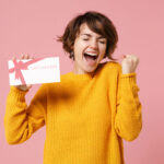 Excited woman holding gift voucher on pink background